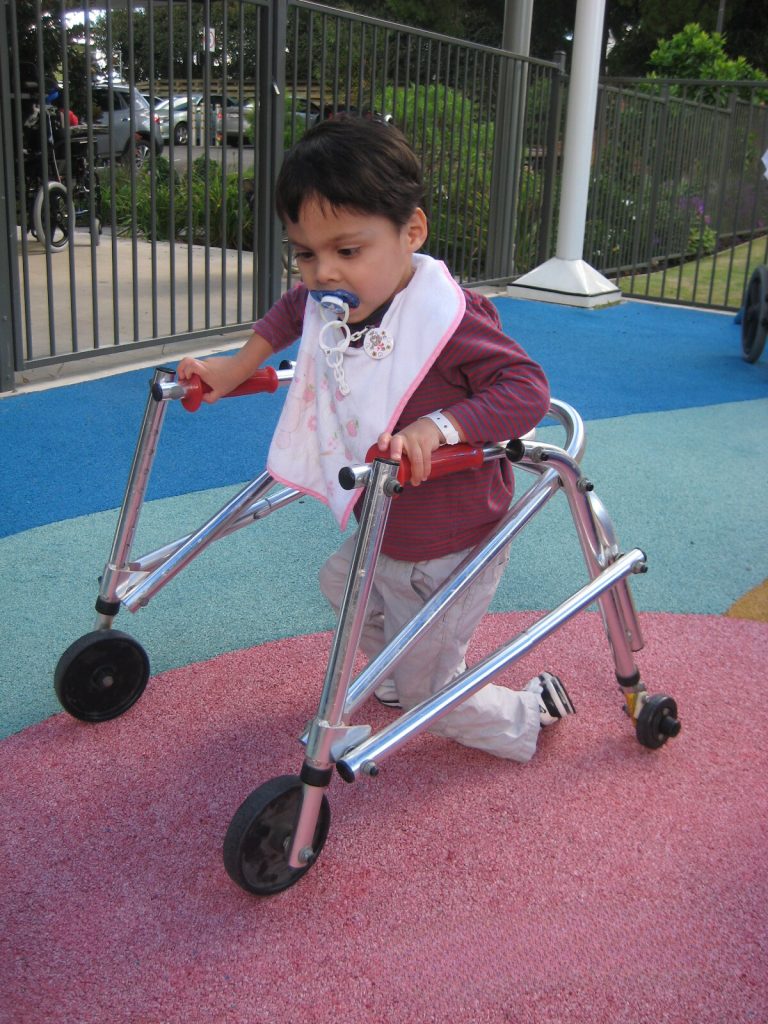 A young toddler using a walking frame in a colourful playground.